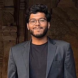 A smiling man wearing glasses and a grey blazer looks directly ahead.